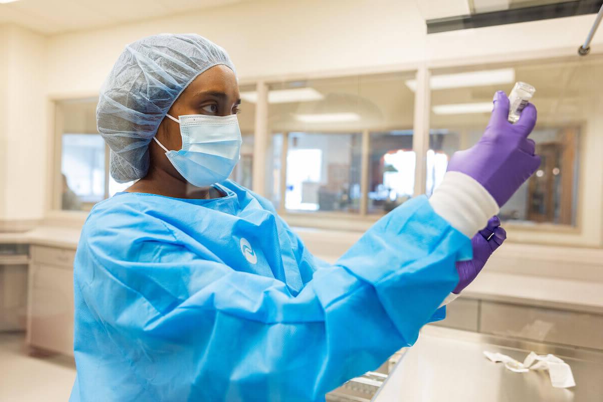 全球最大的博彩平台. Dunn School of 药店 student in blue scrubs, gloves, cap and mask, holding a vial.